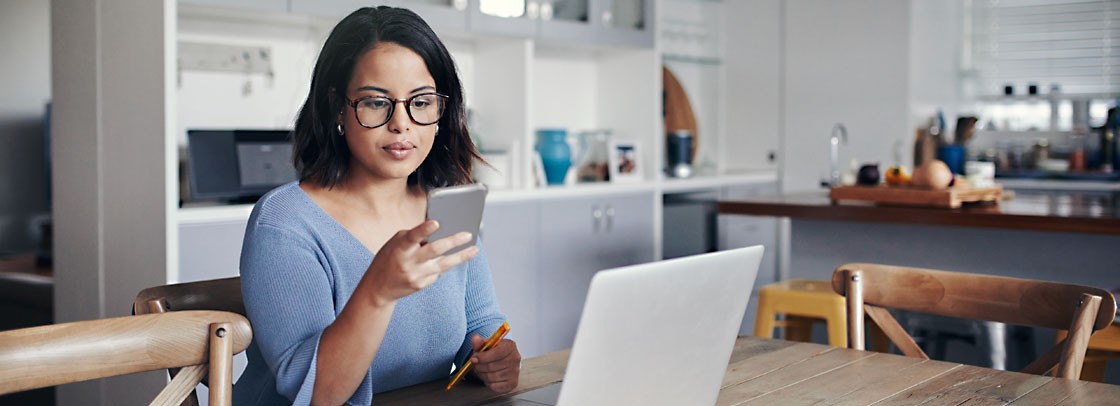 Young lady at table looking at phone and her laptop is on the table.