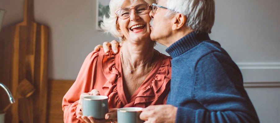 Senior couple kissing in kitchen holding coffee cups