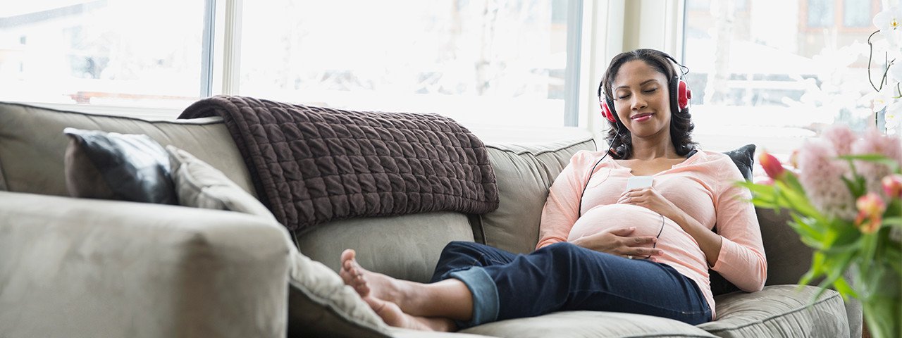 A pregnant woman listens to headphones on the sofa
