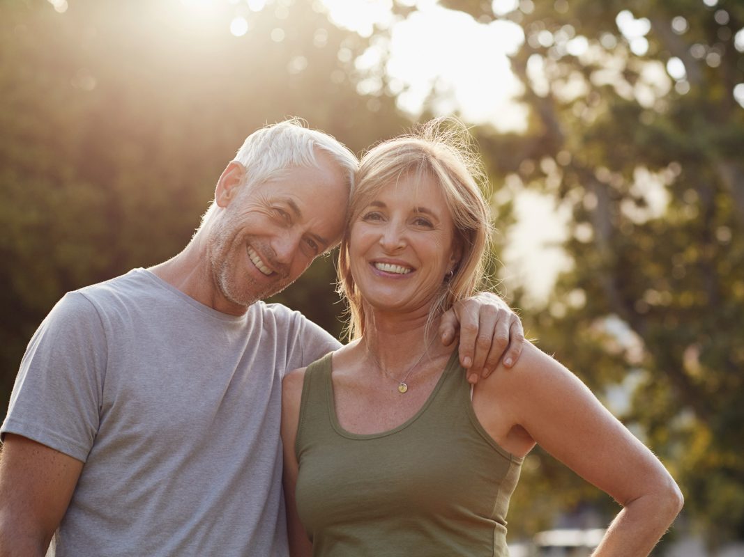 An older couple with their arms around each other smiling with the sun behind them.